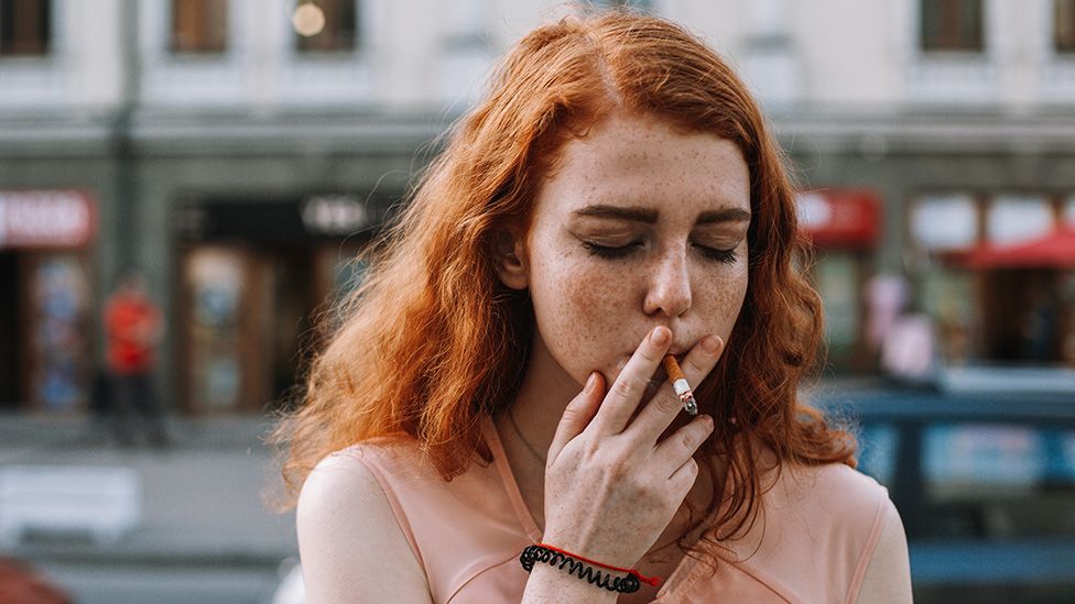 A woman with red hair smoking a cigarette on the street