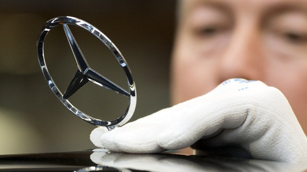 An employee wearing white gloves attaches the Mercedes-Benz star ornament to the front of an S-Class car