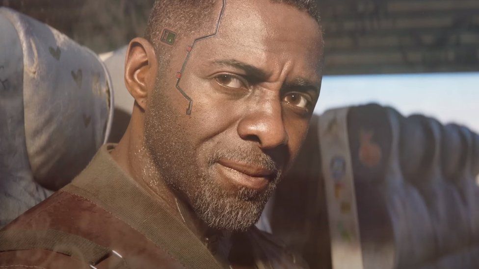 A computer-generated Idris Elba in close-up, sitting in the window seat of a train carriage. His short, stubbly beard is illuminated by sunlight from outside and he has a serious, thoughtful expression. Some sort of wire/track runs from his temple to his cheek, giving him a futuristic appearance.