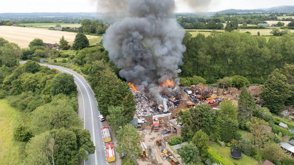 Drone shot of flames and black smoke coming from John Huntley scrapyard. Fire vehicles can be scene on the site and an adjoining road. The site is surrounded by fields, trees and private gardens