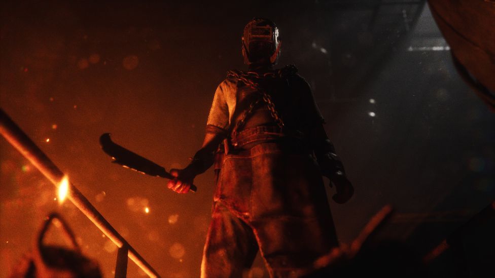 A man wearing a welding mask and leather overalls stands, holding a large knife, in an industrial factory setting. It's dark but illuminated by the glow from a furnace underneath the walkway he's on.