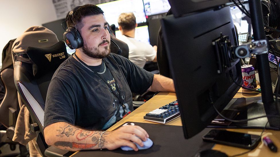 A young man with heavily tattooed arms sits in front of a computer monitor. His hand rests on a mouse and he wears a large set of over-ear headphones. He looks deep in concentration but calm.