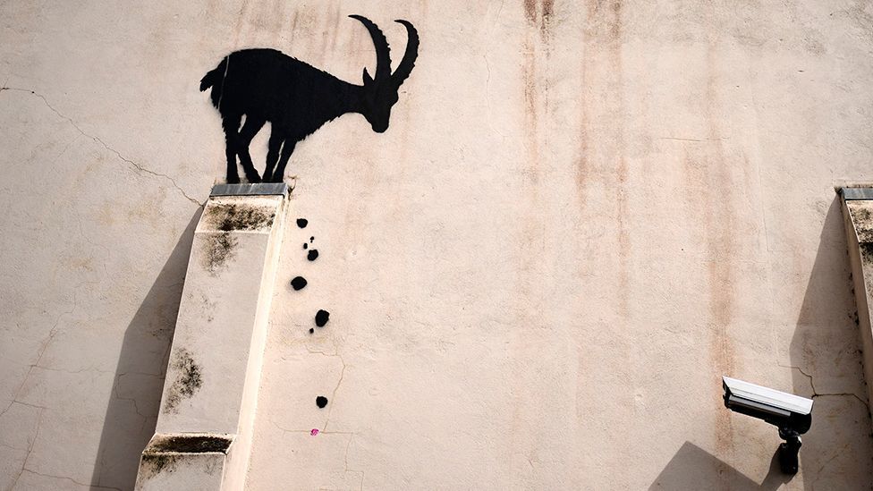 The black silhouette of a goat stands on the external structural support of a building with falling painted black rocks, and a CCTV camera points in its direction.