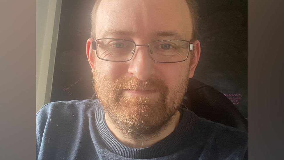 Chris Whitaker, with short fair hair and a close cropped beard, wearing rectangular steel-rimmed glasses, and a grey jumper, smiling at the camera