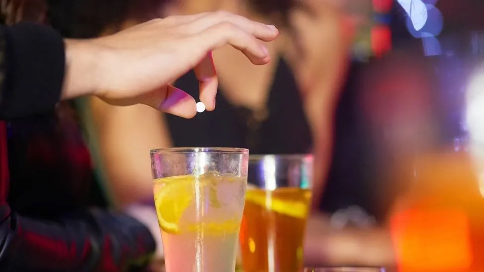 Two glasses sitting on a bar, with a hand above one of them about to drop a tablet into one of the drinks