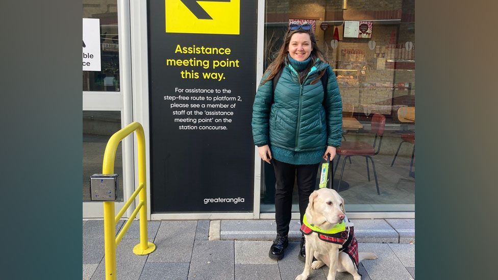 A smiling Samantha Leftwich in a zipped up dark green puffer jacket with a green turtleneck jumper underneath, and black trousers and military style boots. She is holding the neon lead of her her guide dog Lizzie who is wearing a tartan blanket and looking out into the distance 