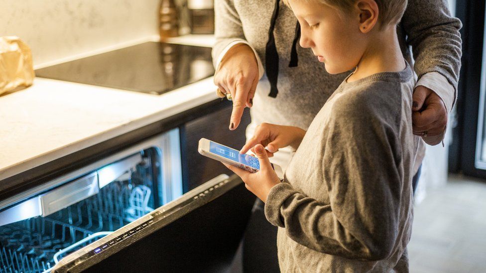 Мальчик учится у матери, используя мобильное приложение вместо посудомоечной машины на кухне в умном доме.