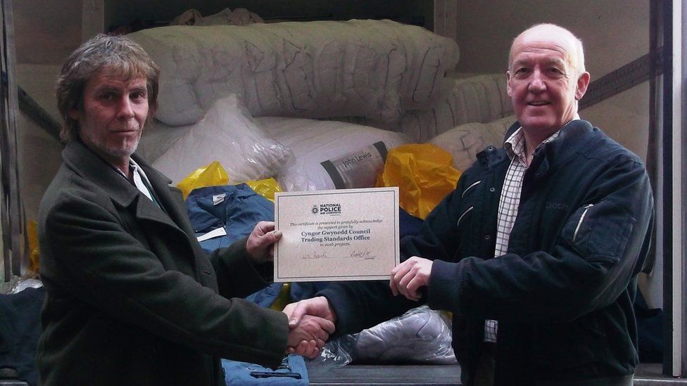 John Eden Jones from Gwynedd Council's Trading Standards Service handing over the counterfeit clothing to David Scott from the National Police Aid Convoy charity