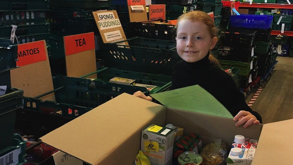 Luna at a foodbank. She is stood in a warehouse near a box of food and is smiling at the camera