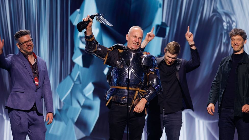 Four men stand on a stage smiling and celebrating. One, dressed in a suit of armour, holds an awards statue - a winged figure with head thrown back as if about to take flight - above his head with one hand. A man to his left in a purple suit smiles and waves, and another immediately behind the armour-wearer has both fingers pointing upwards in celebration. The fourth man, to the right of the shot, looks very pleased but is more restrained.