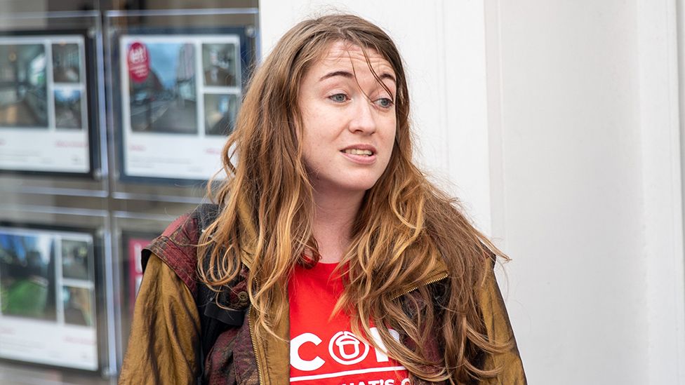 A woman with long, reddish-brown hair stands outside an estate agent's window that's filled with photos and information about properties. She's wearing a brown jacket over a red t-shirt with Acorn written on it in bold white letters. The O has a graphic of an acorn inside it.