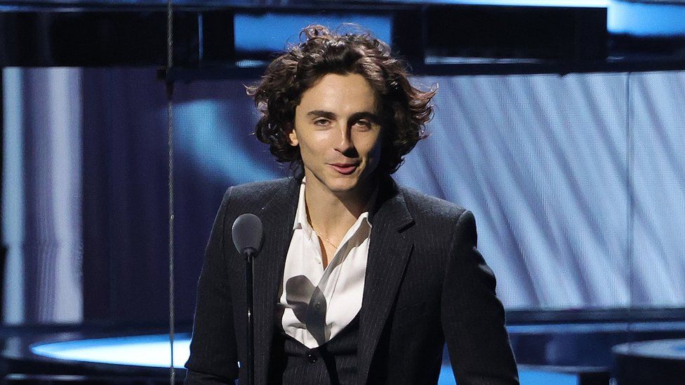 Timothée Chalamet wears a navy three-piece suit with a white shirt open at the neck. He's standing in front of a light blue patterned stage backdrop and speaking into a microphone on a thin stand. He looks relaxed and is smiling as his medium-length black hair cascades down, framing his face.