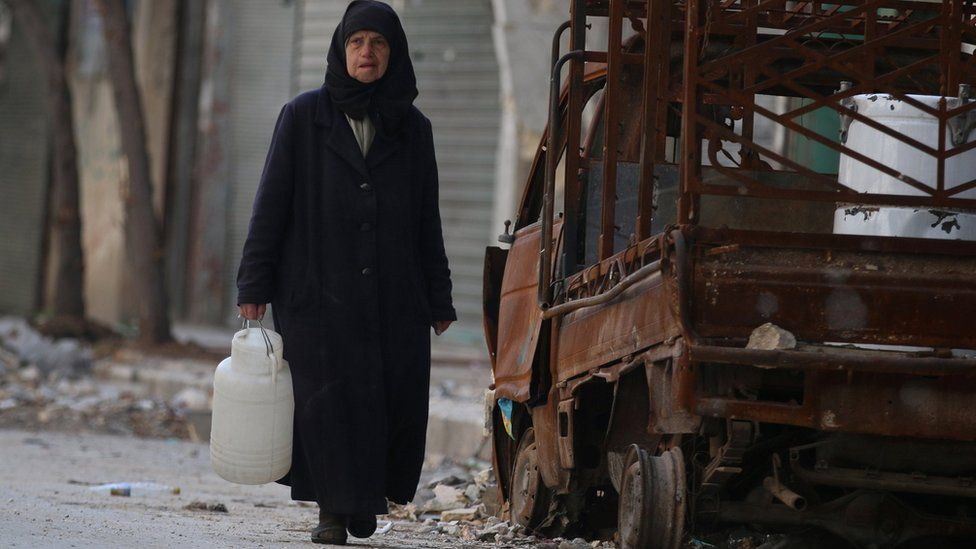 A woman carries a container of water as she walks past a burnt pick-up truck in a rebel-held area of Aleppo's Old City (14 November 2016)