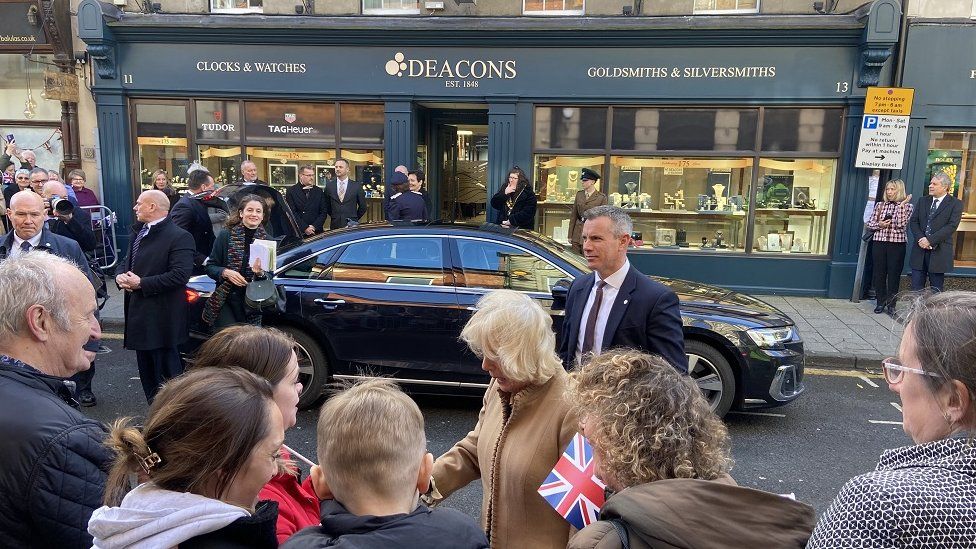 The Queen meets the public in front of a car, protection officers and Deacons shop front
