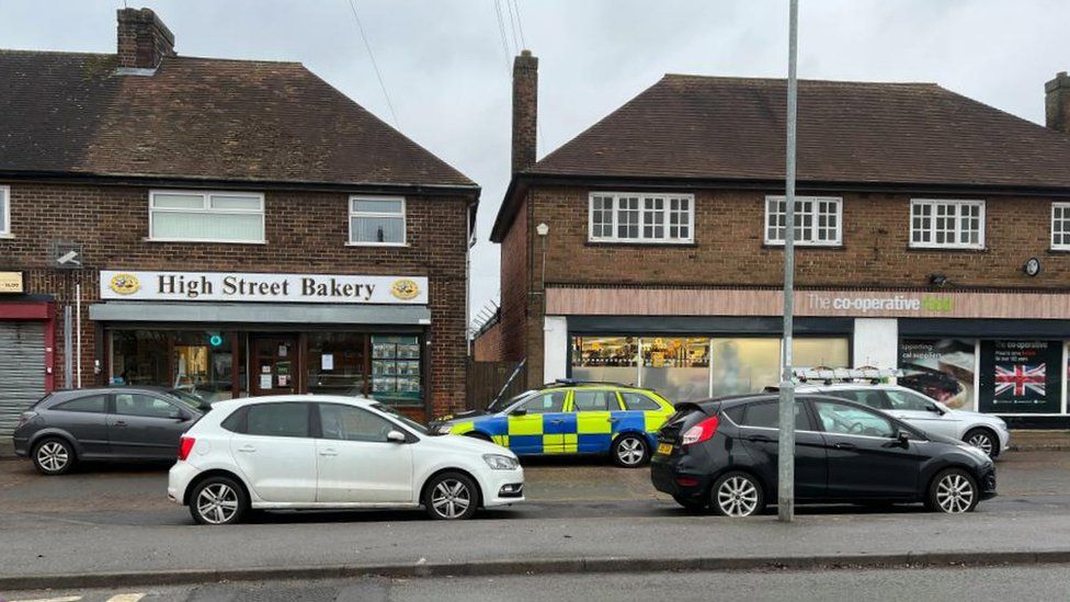 Parade of shops including bakery and Co-op with police car parked alongside a gate to a path between two shops
