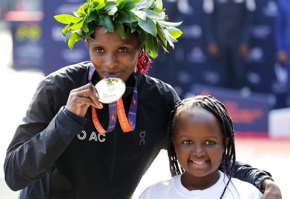 A woman holds a medal and her young daughter smiles next to her.