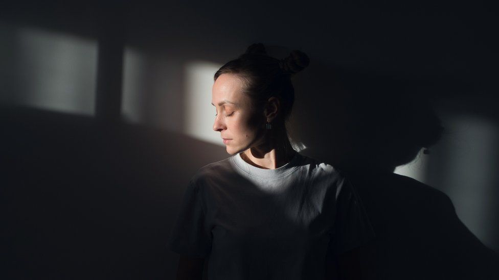 A lonely Young Sad Calm Girl Or Woman stands against a white wall, in a psychiatric clinic or at home, in the rays of sunlight, with her eyes closed. The Concept of Depression from Illness and Quarantine, Fatigue and Impotence. A copy of the space. - stock photo