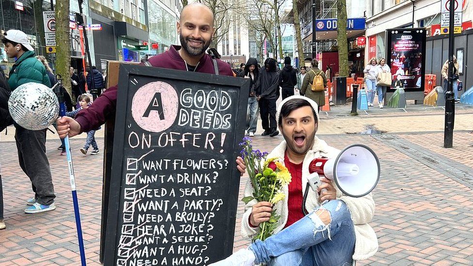 Asian Network's Haroon Rashid - a bald man with dark beard - stands in a busy High Street wearing a chalk board with a list of good deeds on it. They are "Want Flowers? Want a Drink? Need a Seat? Want a Party? Need a Brolly? Hear a Joke? Want a Selfie? Want a Hug?" Shabaz Says sits next to him, holding flowers in one hand and a megaphone in the other. He's pulling a mock-surprised/shocked expression.