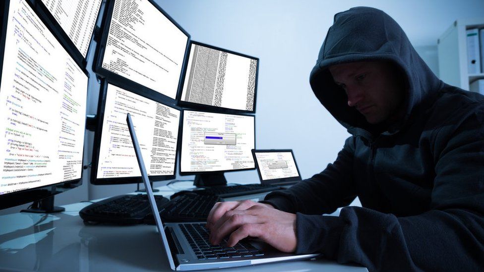 Hooded hacker on laptop i nfront of bank of desktop monitors
