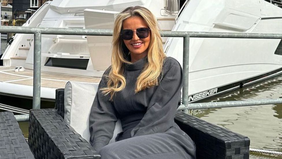 A young white woman with long blonde hair sits in front of a yacht. She's on an outdoor armchair, wearing sunglasses and a loose, polo-necked tracksuit in a luxurious dark grey colour.