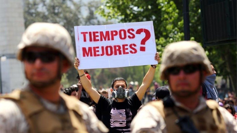 A demonstrator holds a sign that reads "Better times?" as soldiers stand guard at Plaza Italia square in Santiago, Chile, on October 21, 2019.