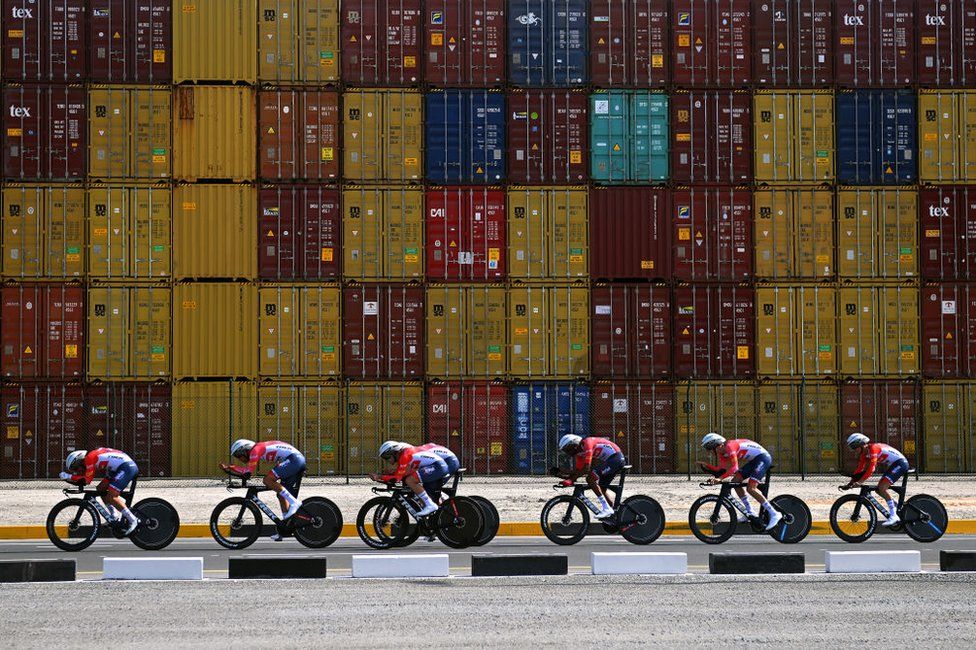 Cyclists race past stacked shipping containers.