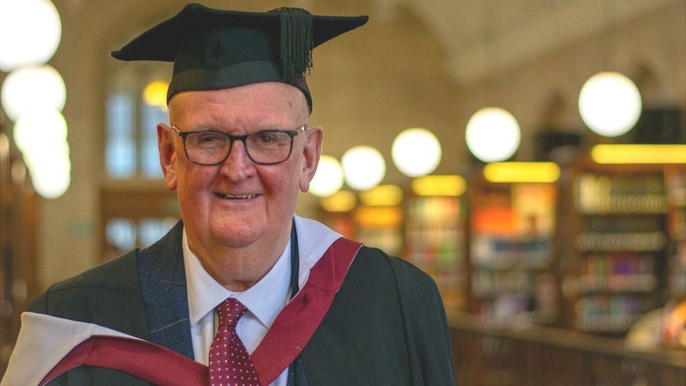 Paul Deal standing in a library wearing his graduation robes and looking at the camera