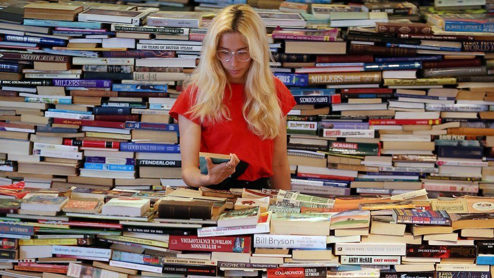 A blonde woman stands in the middle of a huge pile of books.