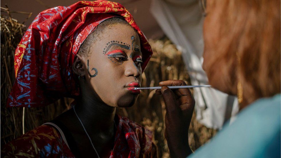 A woman having her face painted. She is wearing a traditional head dress and has on red face paint on her eyelids and red paint on her lips