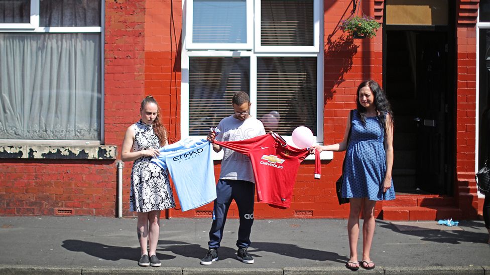 Three people hold up a Manchester United shirt and a Manchester City shirt