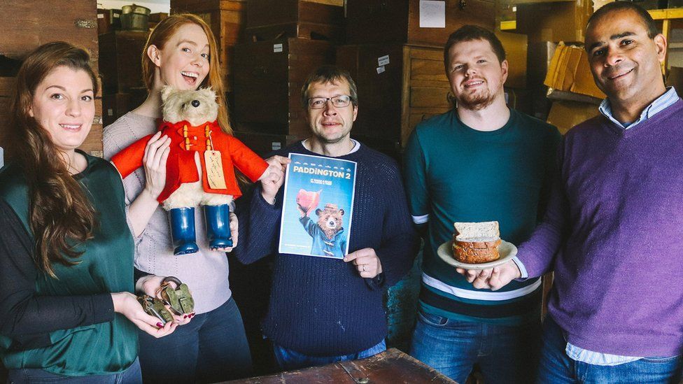 Two women and three men holding padlocks, a Paddington bear, a movie poster for Paddington 2 and a plate of marmalade sandwiches