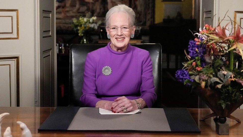 Queen Margrethe II, wearing a purple dress, smiles as she sits behind a desk
