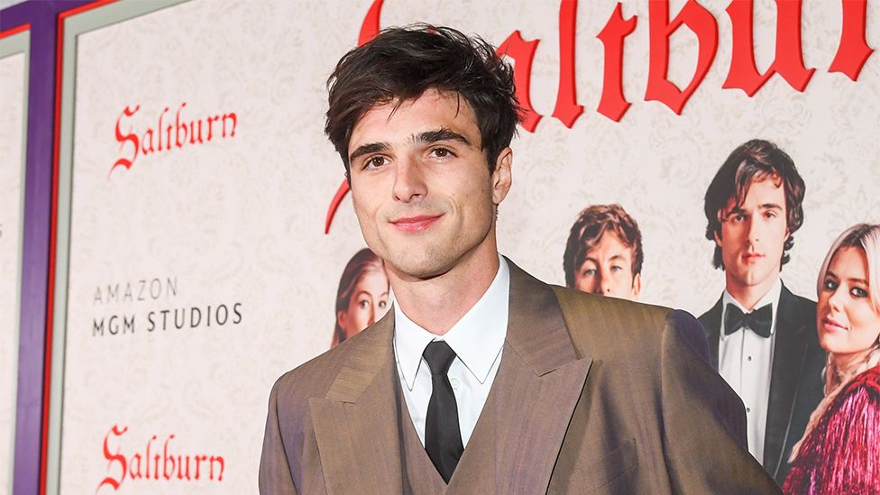 Jacob Elordi, wearing a brown suit with white shirt and black tie, looking to the side of the camera. The background is a poster of "Saltburn" with images of the cast and red writing on a white background.