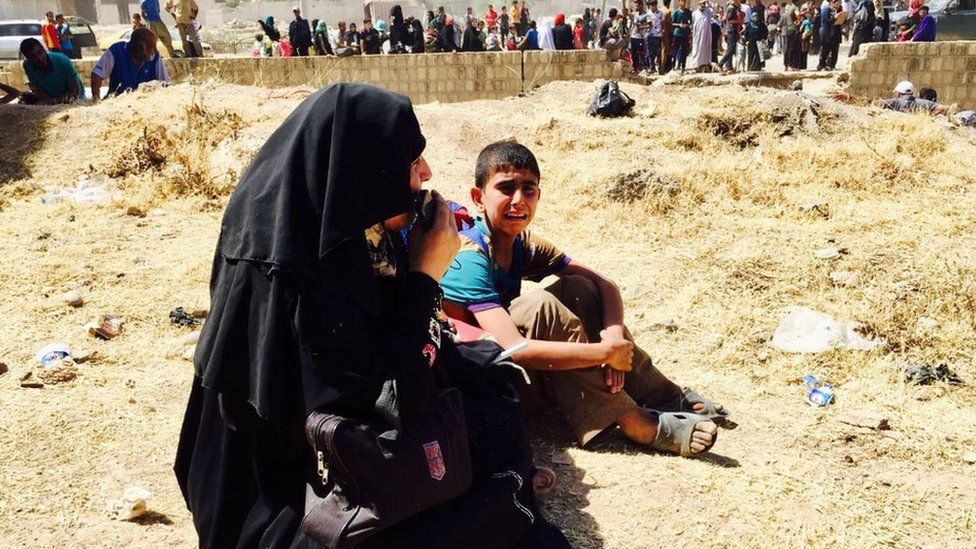 A young boy cries as he sits on the ground with a woman, in front of dozens of other civilians
