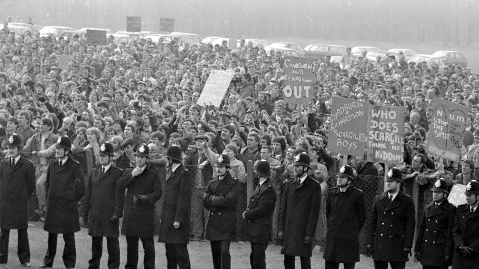 A line of uniformed police officers stand in front of a large crowd of protesting miners