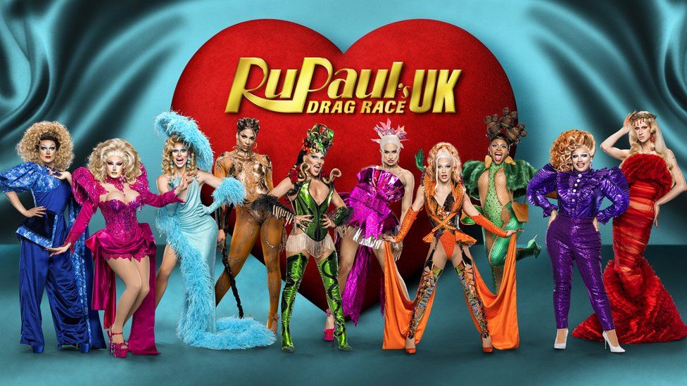Promotional picture for the new series of Drag Race UK. The 10 drag queens are stood side by side posing with their hands on their hips or by their sides or on their heads. They're wearing a variety of brightly coloured dresses ranging from blue to pink to gold to green to orange to purple and red. The floor and background is light blue and behind them is a giant red heart with the words "RuPaul's drag race UK" in gold writing on it.