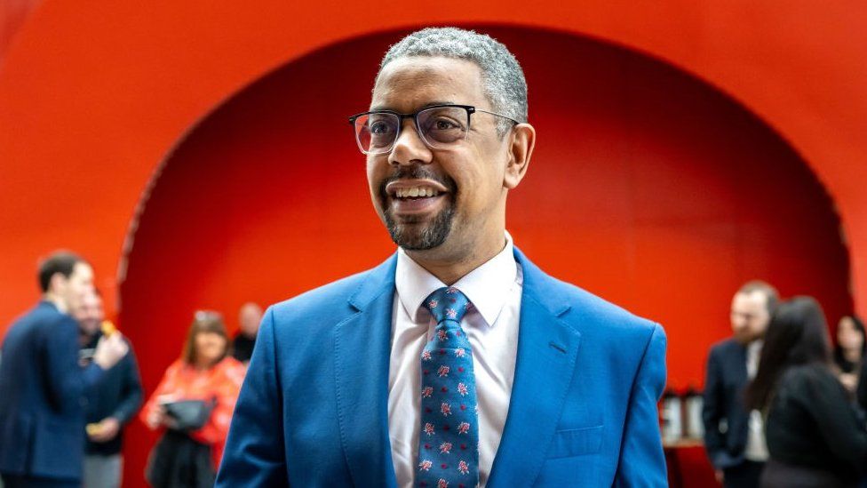 Vaughan Gething, wearing a blue suit, smiles at the camera against a red background