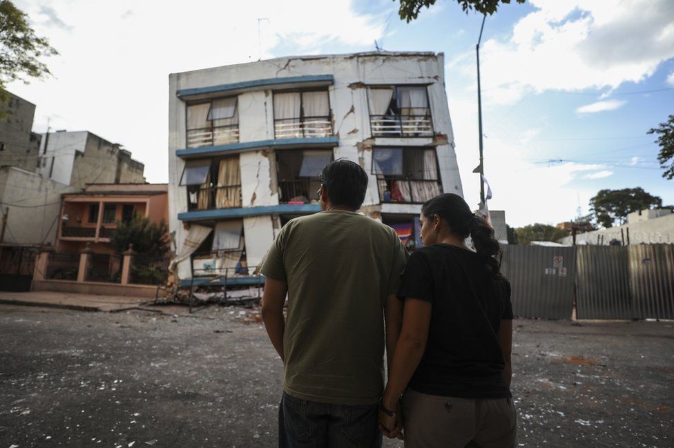 Man and woman stood in front of a damaged building. What used to be a five-storey block has lost its bottom two levels entirely.