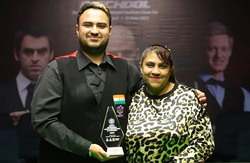 A smartly dressed young Asian man stands with his arm around a woman - his mum - in front of a background with the faces of famous snooker players on it. He wears a black shirt and waistcoat - the waistcoat has the orange, white and green Indian flag embroidered on the left breast. His mum's long hair is tied back, and she's wearing a leopard print top. They're both smiling, and the young man's free hand cradles a glass, diamond-shaped award of some sort.