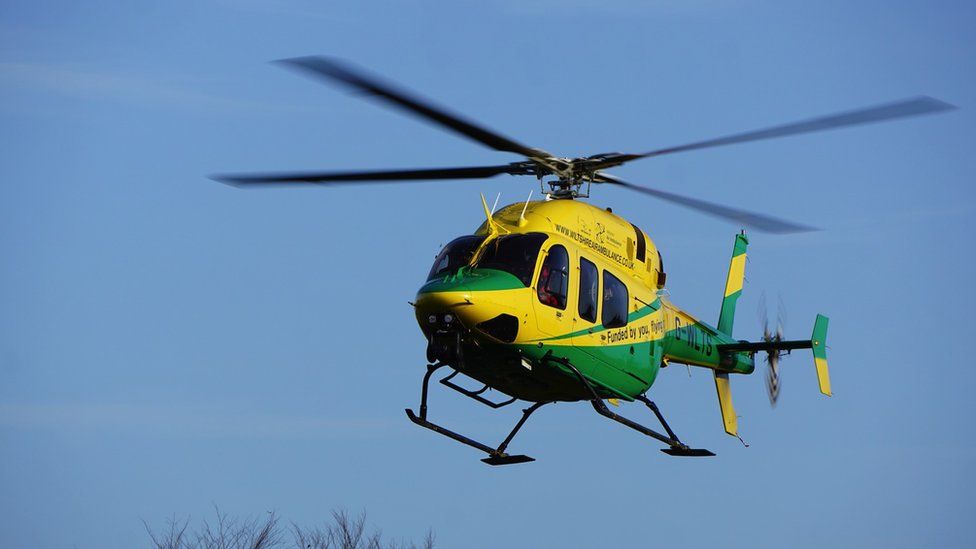 Image of a Wiltshire Air Ambulance helicopter in the sky. It is yellow and green.
