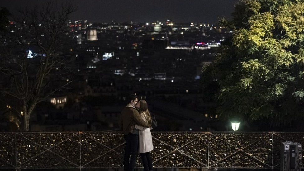 A couple wearing protective face masks embrace, in front of the Paris skyline seen from Montmartre at nightfall, just hours before a city-wide night time curfew goes into effect in Paris, France, 16 October 2020