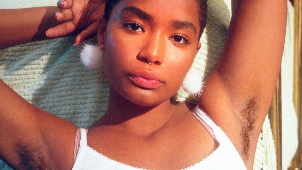 A young black woman poses with her arms above her head, showing her armpit hair
