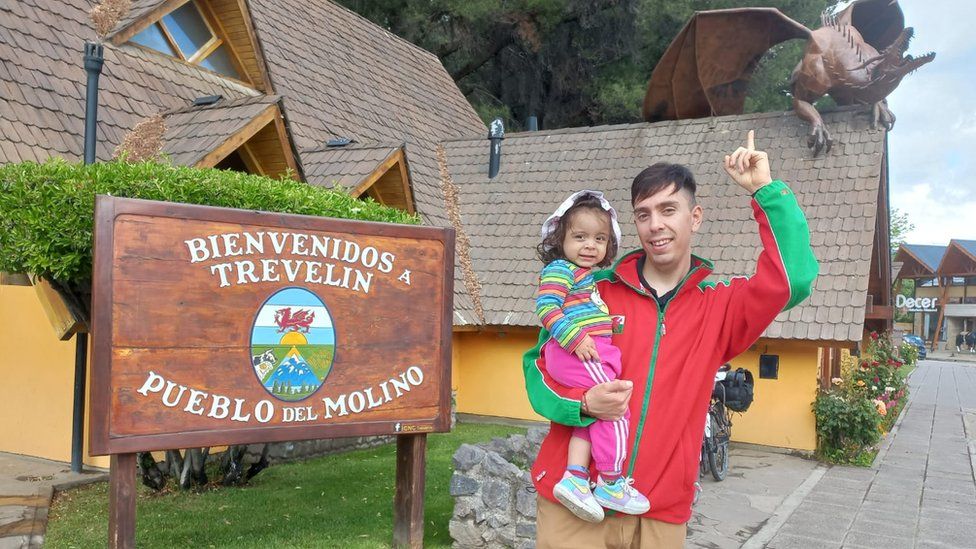 A photo of David and his daughter next to a Welcome to Trevelin sign. The Trevelin flag is central on the sign with the Argentinian colours and a red dragon in the middle.