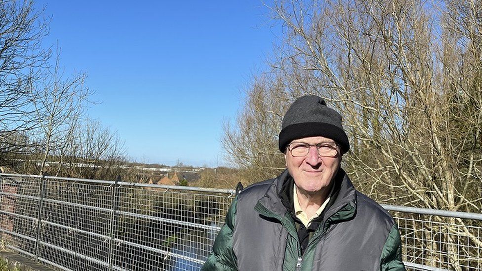 Road Hacker looks at the camera while standing on a bridge over the canal, wearing a puffy winter coat, glasses and woolly hat