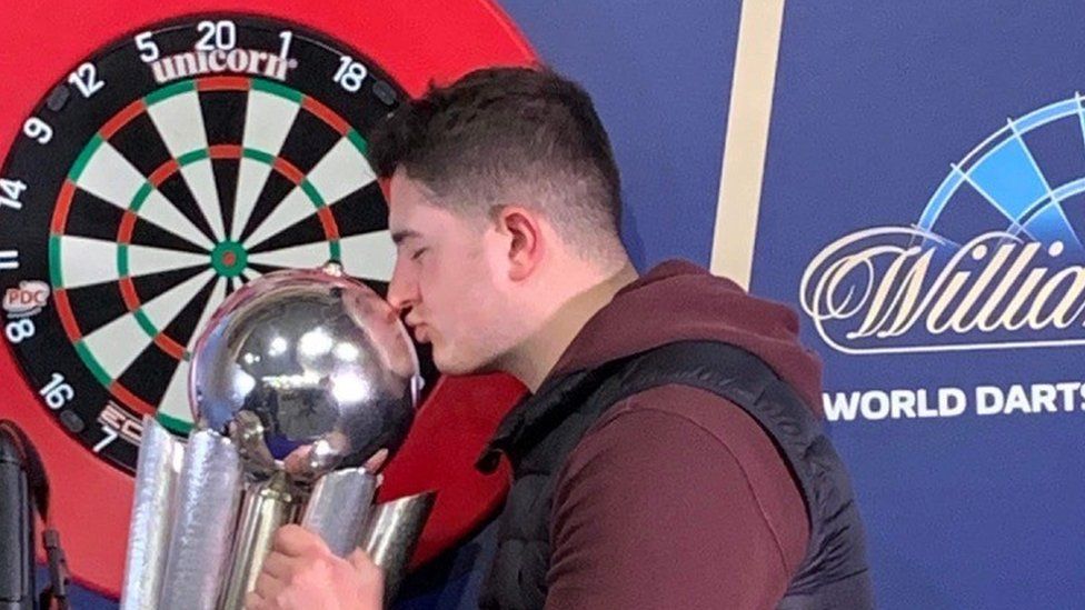 A man wearing a red hoodie and a navy jacket kisses a silver trophy in front of a darts board