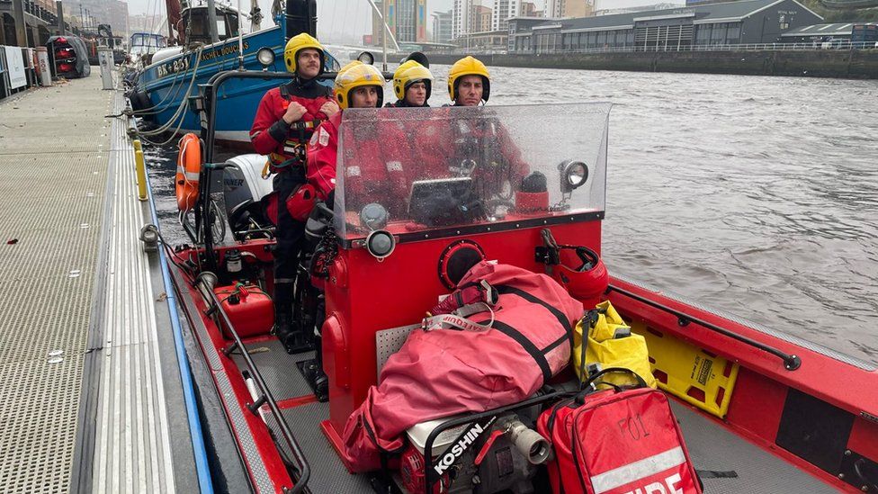 Four firefighters in red water suits and yellow helmets stand aboard a fire service boat on the Tyne