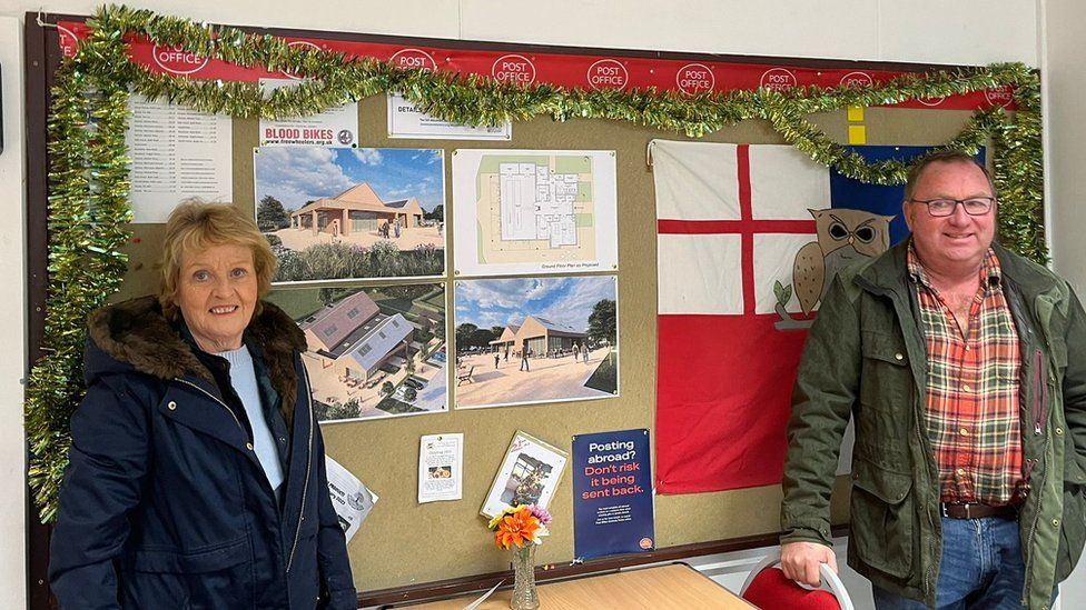 Sue standing on the left and Jim on the right in front of a noticeboard detailing plans of the proposed new building