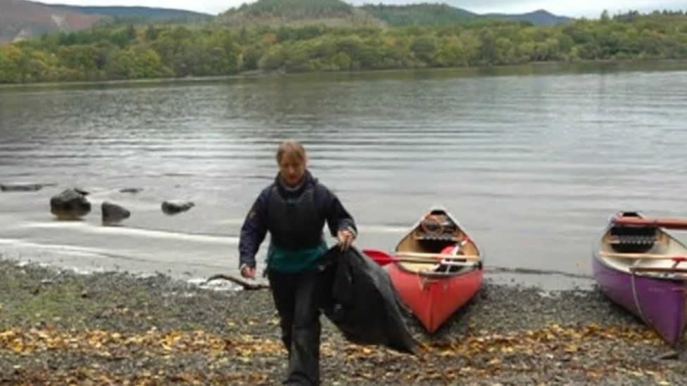 Volunteer on island shore carrying a black refuse sack with canoes at waters edge
