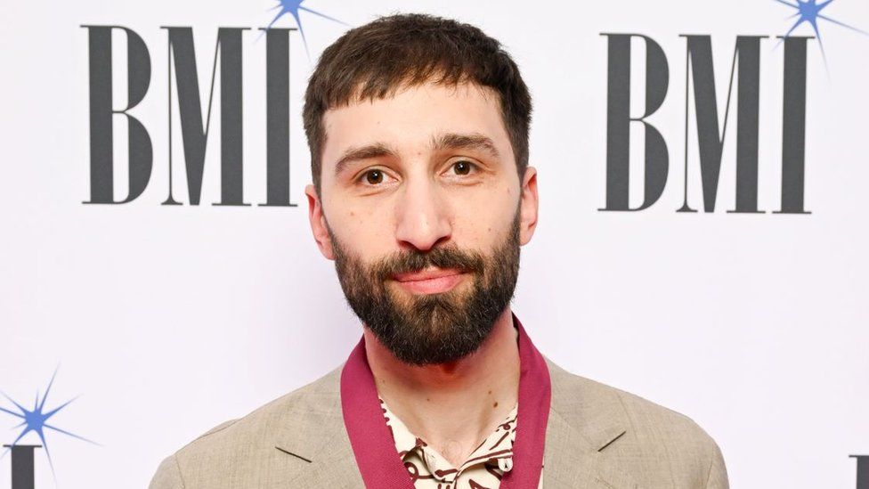 Linden Jay at the BMI Awards. Linden, a young man, has short brown hair and a short dark beard. He has brown eyes and looks at the camera with a slight smile. He wears a beige suit over a cream coloured shirt with a maroon line pattern and a red scarf. He is pictured in front of a white back drop with the BMI logo printed on it.