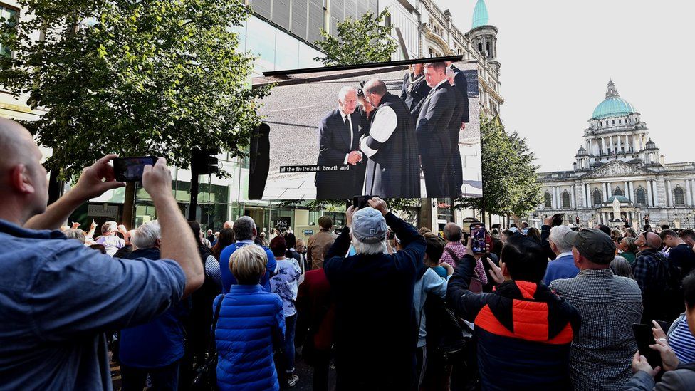 A crowd of people in Belfast city centre watch a large screen broadcasting the arrival of King Charles at St Anne's Cathedral